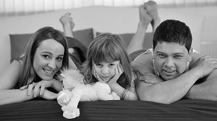 families greyscale family of 3 laying on their stomach with feet at the head of the bed, the daughter has a plush toy with her and they are all smiling at the camera